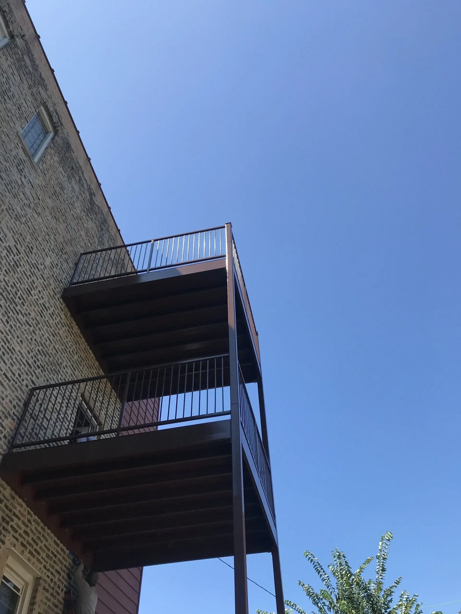 Gallery. Two-story exterior deck attached to a brick building. Dark brown deck and railing, against a clear blue sky.