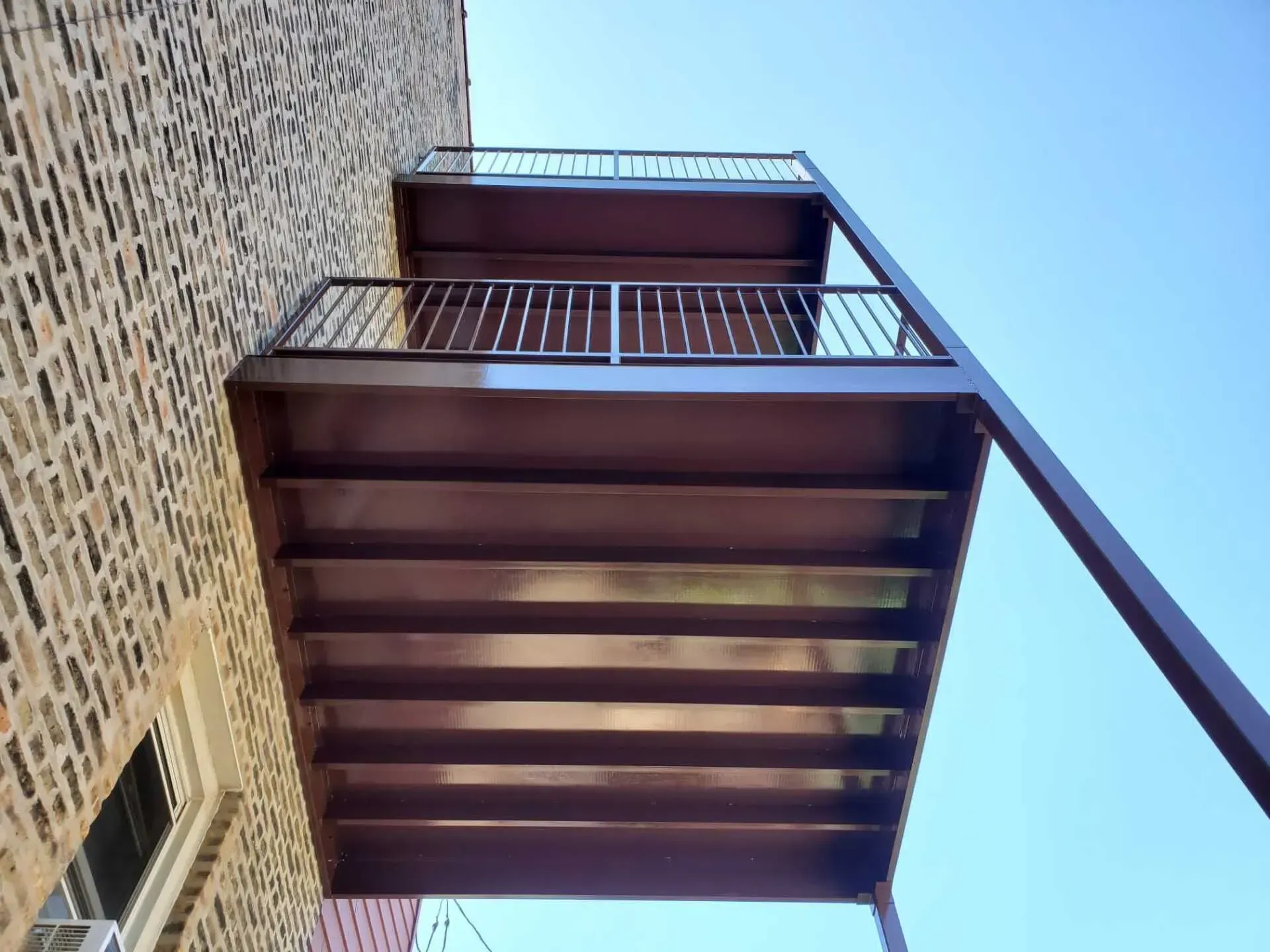 Gallery. Two-story brown metal balcony attached to a brick building, viewed from below against a blue sky.
