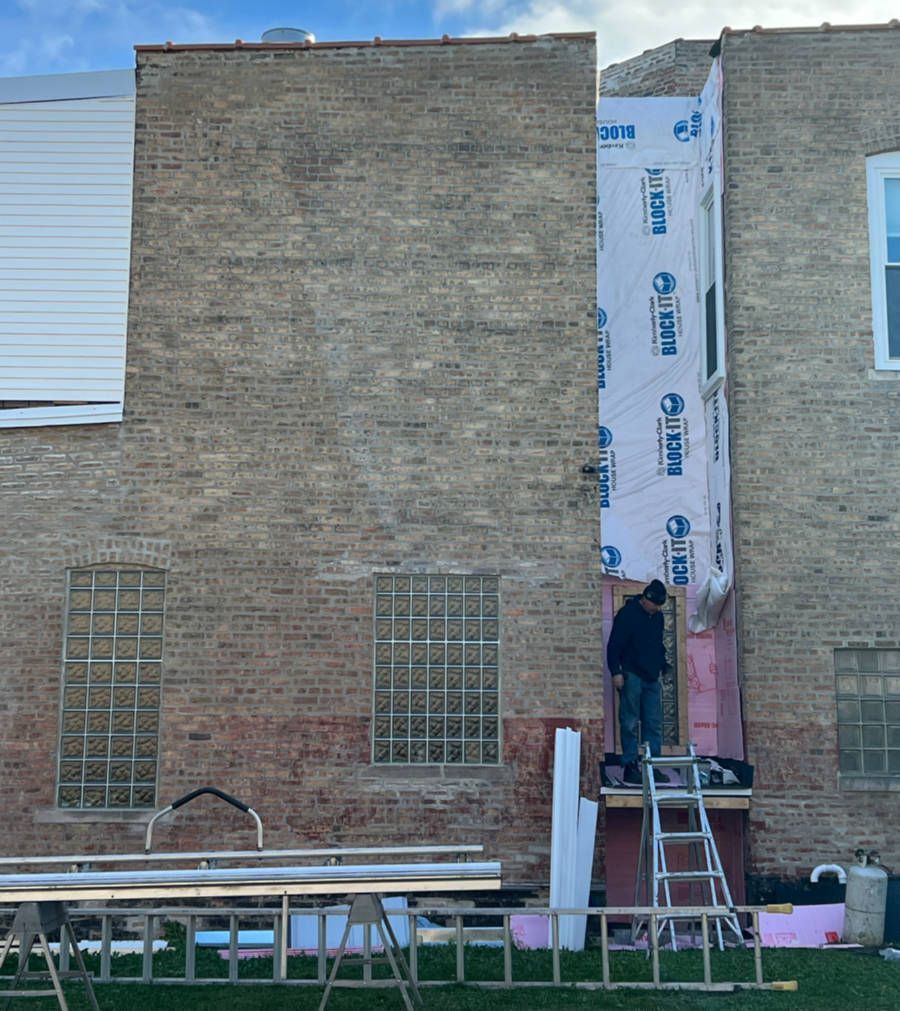 Construction worker on a ladder, working on the side of a brick building. Blue wrap, pink insulation, and tools are visible.