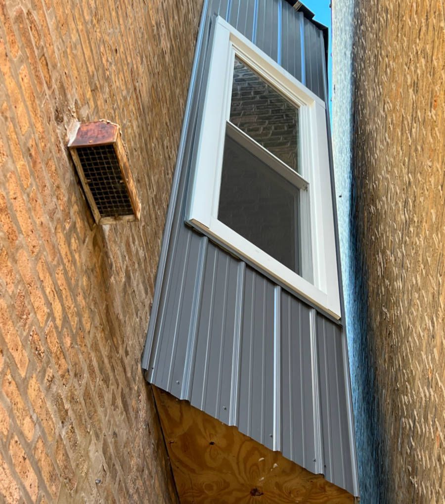 Exterior view of a narrow space between two brick buildings. Gray siding and a white window are installed. A vent is visible.