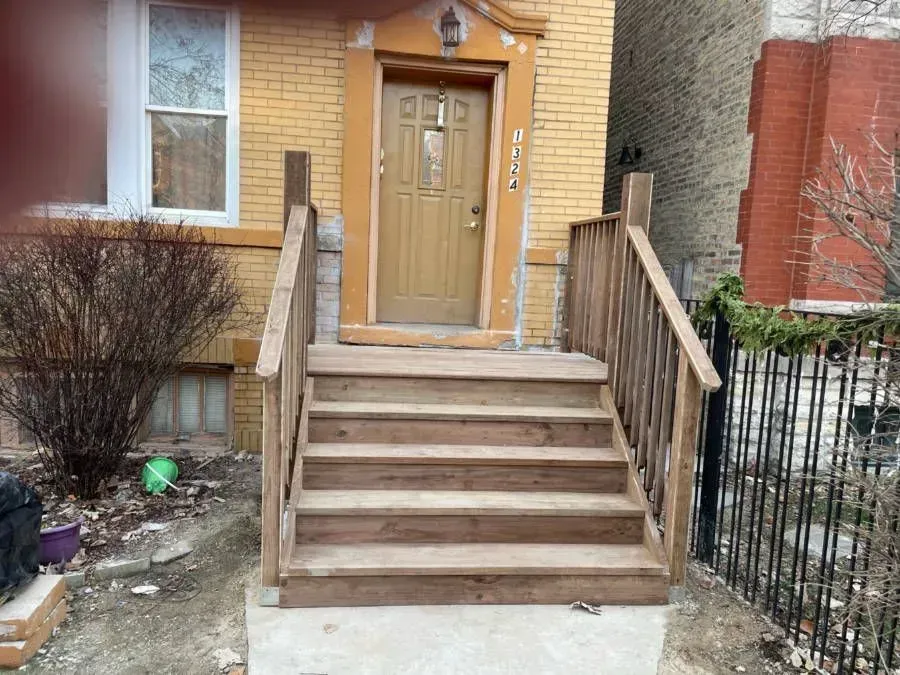 Gallery. Wooden steps leading to a beige door, railing on the side. Yellow brick house, winter setting.