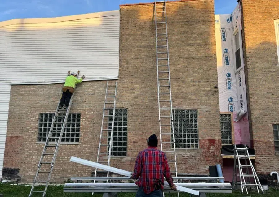 Construction workers on ladders renovating building exterior. Brick, siding, and weather barrier visible.