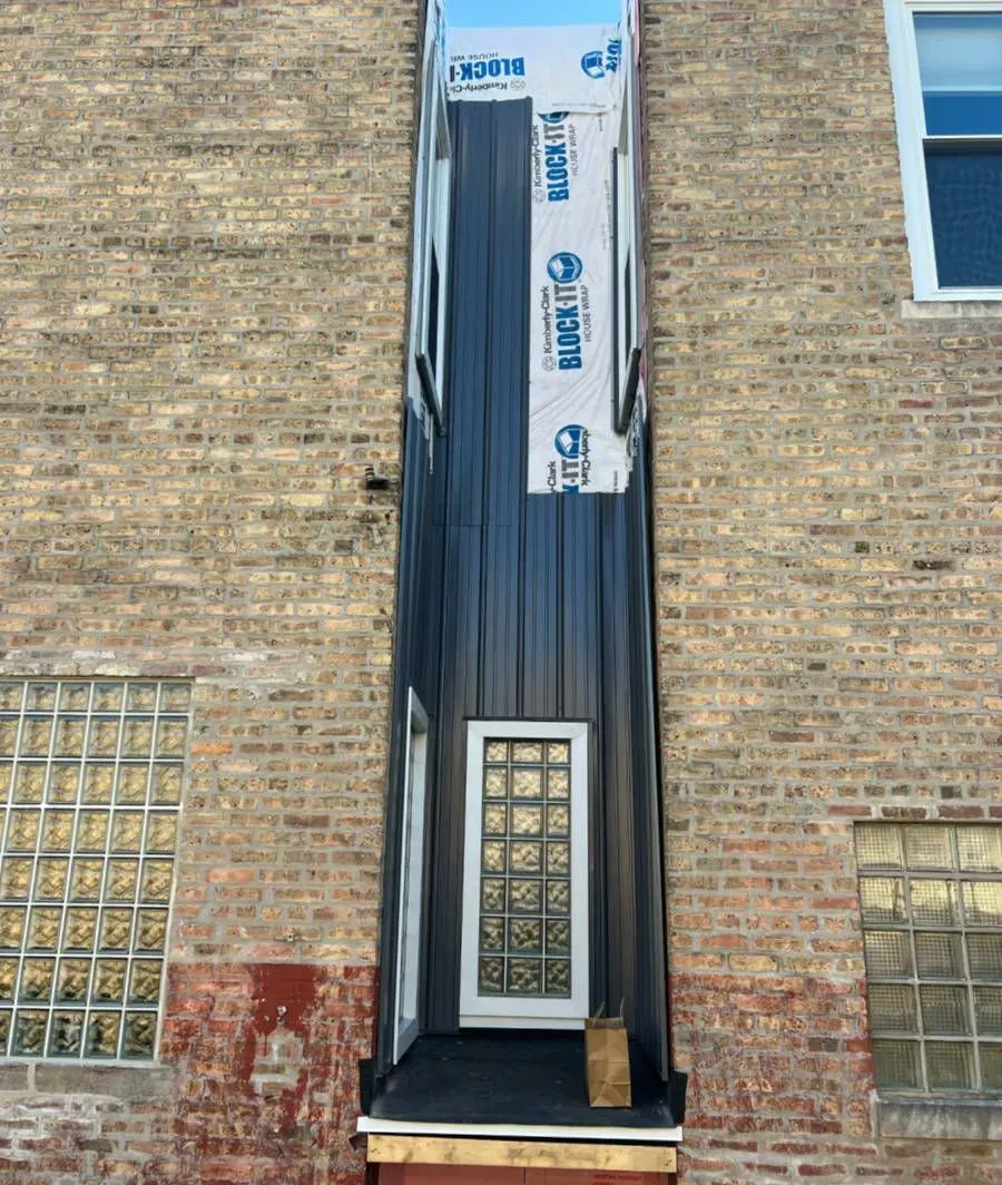 Narrow, dark metal-clad exterior of a building with a door and windows set into a brick wall.