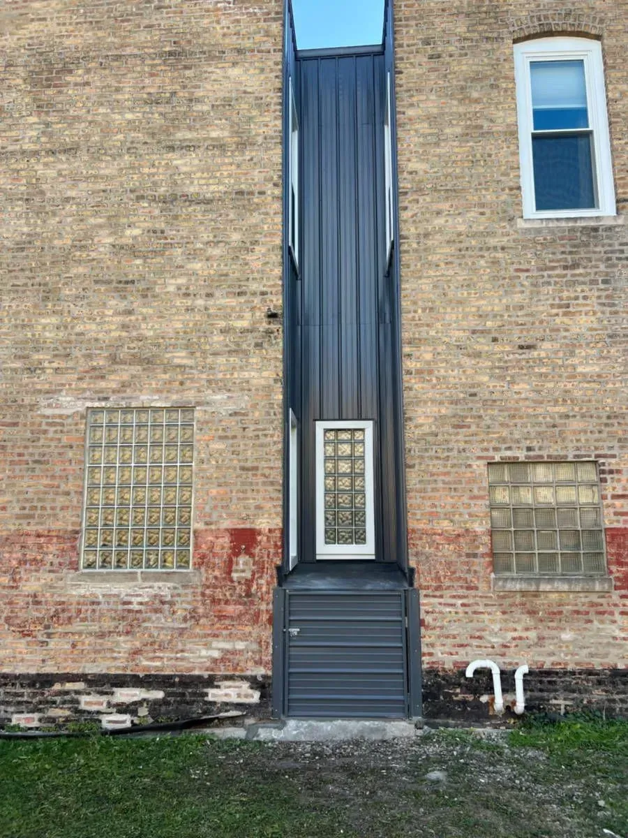 Dark metal elevator shaft on brick building with windows and door.