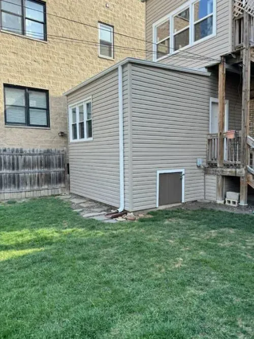 A small beige building with a window and a door in a grassy backyard.
