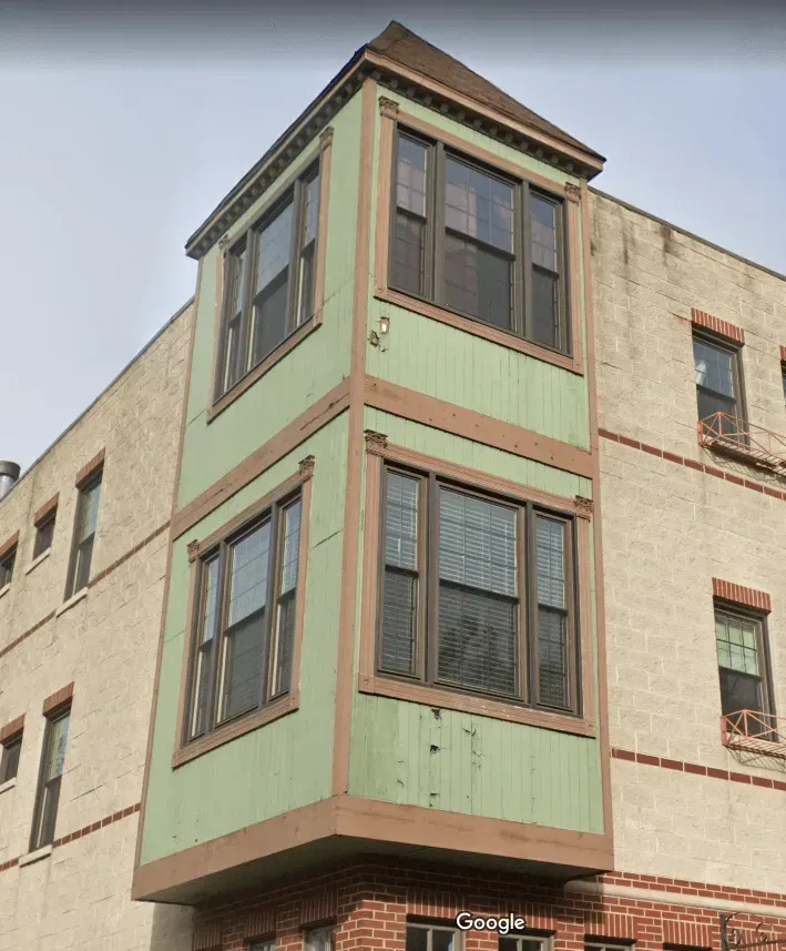 Two-story building corner with green and brown bay windows, tan brick exterior, and a brown roof.