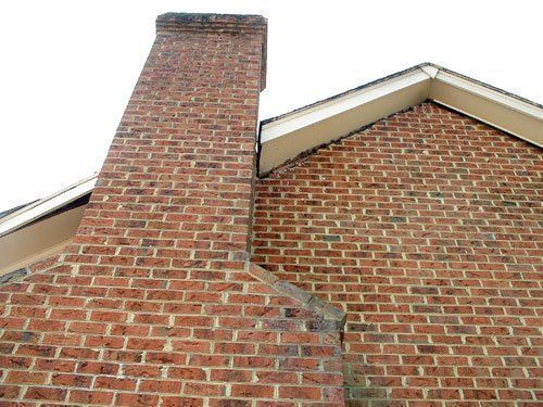 GALLERY. Brick chimney attached to a brick wall of a house; red-orange bricks, white roof trim.
