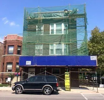 Building with scaffolding and netting, blue awning, black SUV parked in front.
