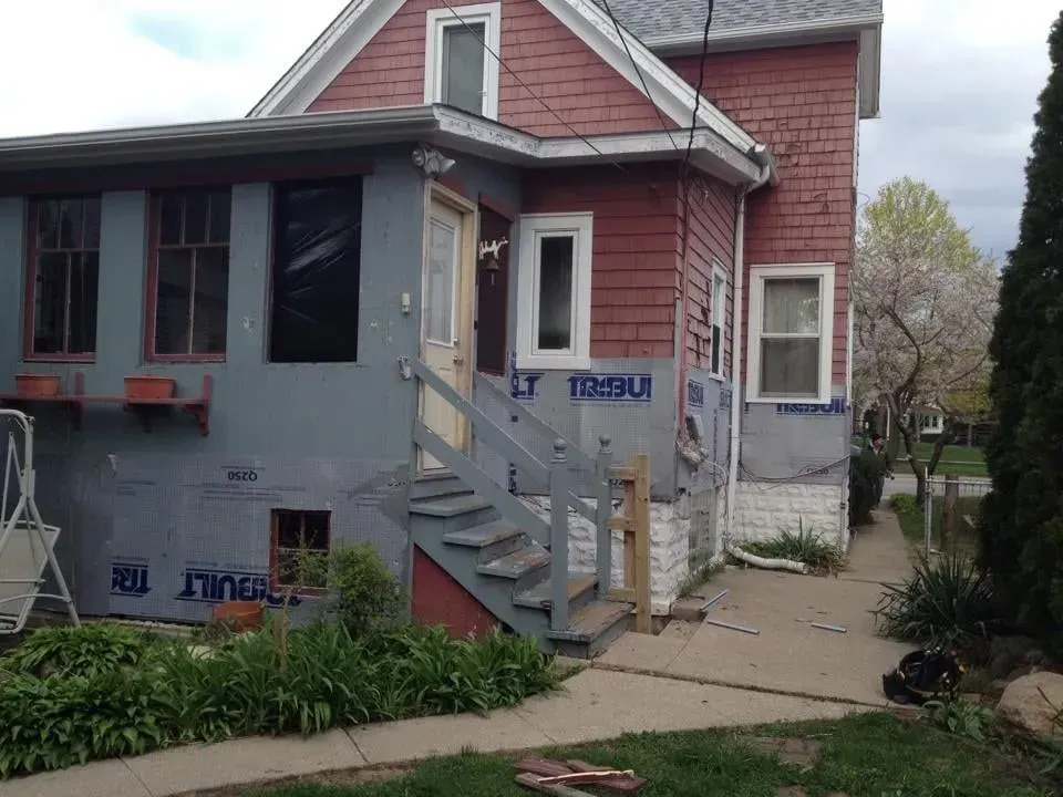 Side view of a house with red siding and gray insulation. A set of stairs leads to a front door.