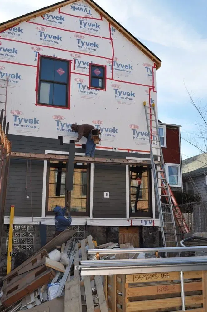 Construction site: building exterior with windows, siding, Tyvek wrap, workers, ladder, and materials.