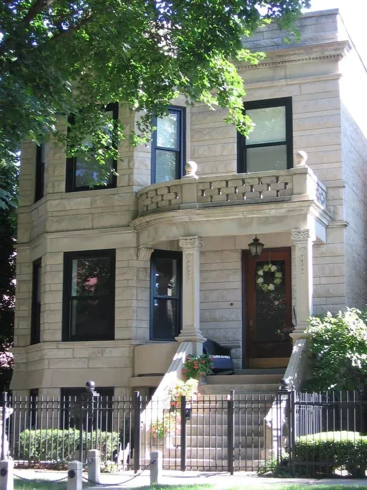 Three-story stone building with a wrought-iron fence, front porch, and black-framed windows.