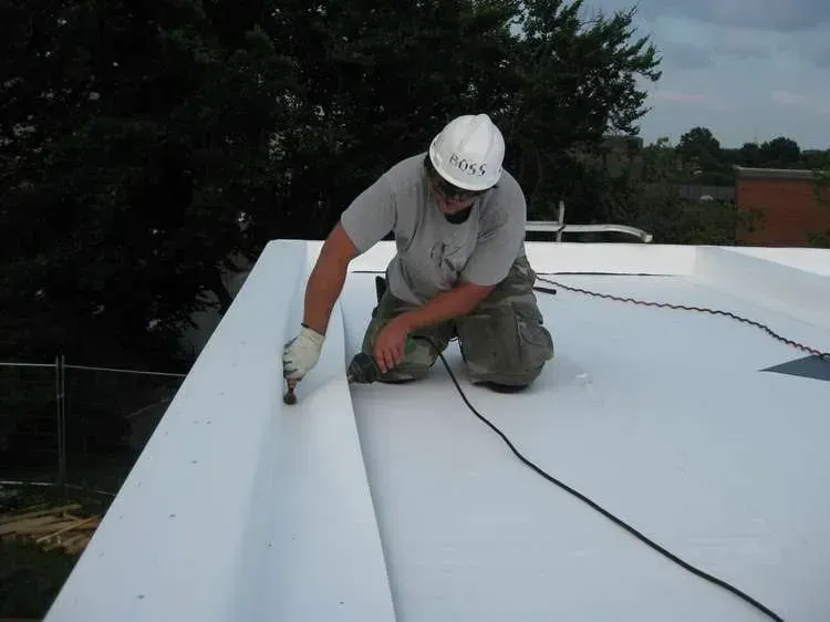 Roofer kneeling, installing white roofing material on a flat roof, wearing a hard hat.