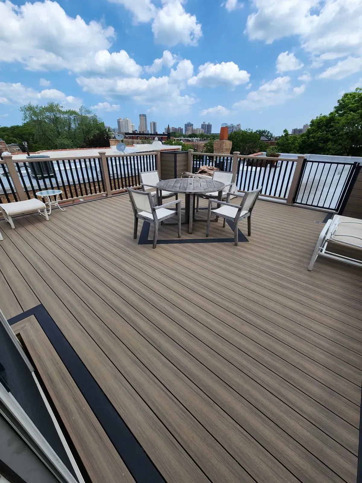 Gallery. Rooftop deck with a round table and chairs. Black railings surround the wooden deck, with cityscape in the distance.