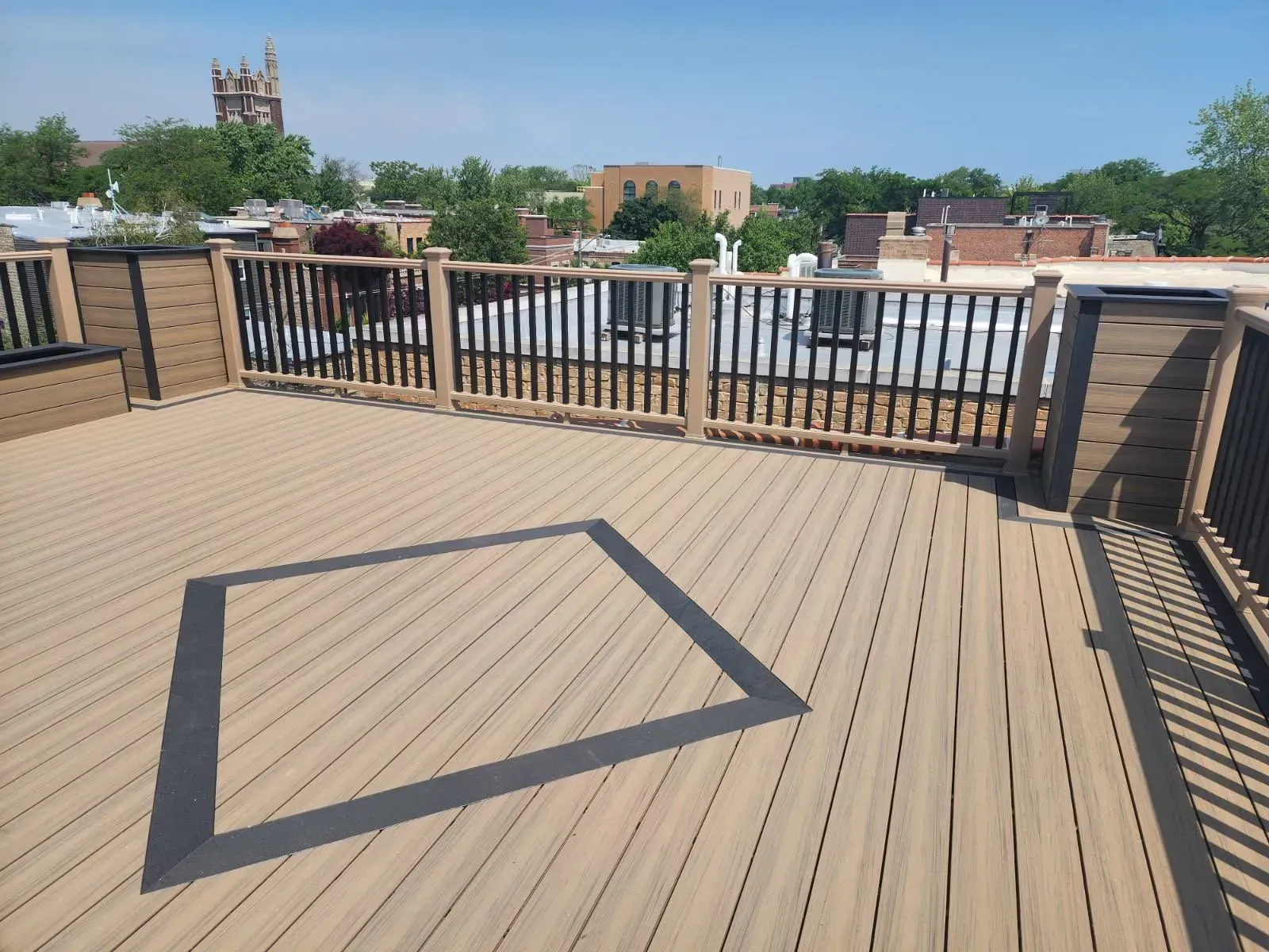Gallery. Rooftop deck with tan decking, black and tan railings, and a square inlay. City buildings in the background.