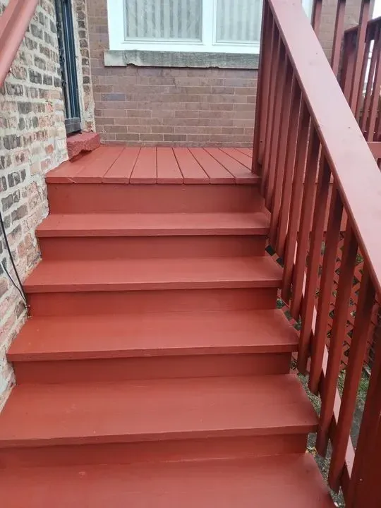 Gallery. Red painted wooden steps leading up to a deck, with a brick wall to the left and railing to the right.