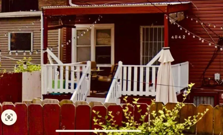 Gallery. Red porch with white railing, partially shaded by an umbrella, behind a red fence, with string lights.