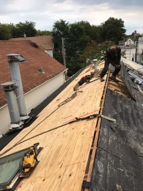 Gallery. Person on roof replacing plywood. Partial view of two buildings, chimney, and trees.
