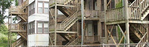 GALLERY. Wooden fire escapes on a multi-story building. Weathered wood, brick facade, and a chain link fence.