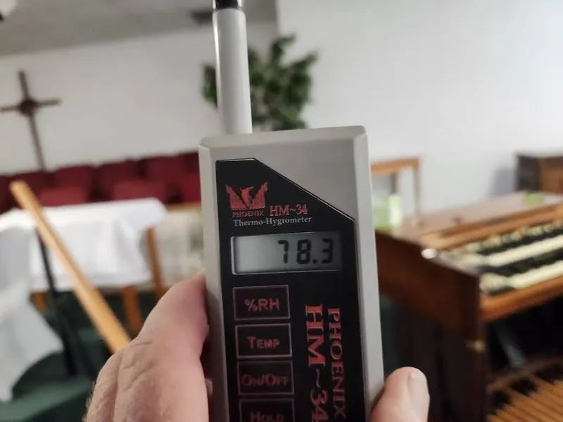 Handheld digital hygrometer in foreground, showing 78.3°F in a room with tables and chairs.