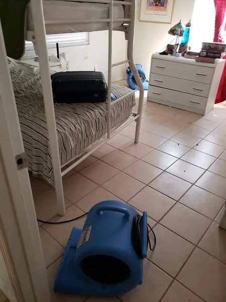 Bedroom with a white bed, black suitcase, dresser, and a blue air mover 
on the tiled floor.