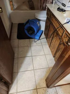 Bathroom floor with a blue air mover fan on tiles beside a toilet and vanity cabinet.