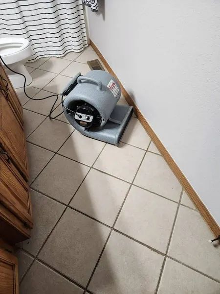 Gray air mover fan on a tiled floor beside a white wall and curtain