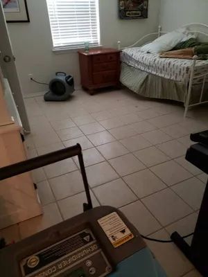 Bedroom with tiled floor, twin bed, nightstand, and a air mover near the window.