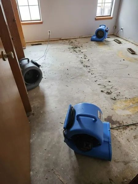 Empty room with water damage, debris on floor, and blue air mover fans drying the carpet.