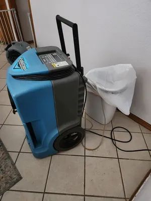Blue dehumidifier on tiled floor beside a white-covered chair and tangled cord in a room