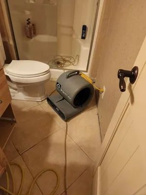 Bathroom with toilet and an air mover on the floor, cords and hose scattered across the tile.