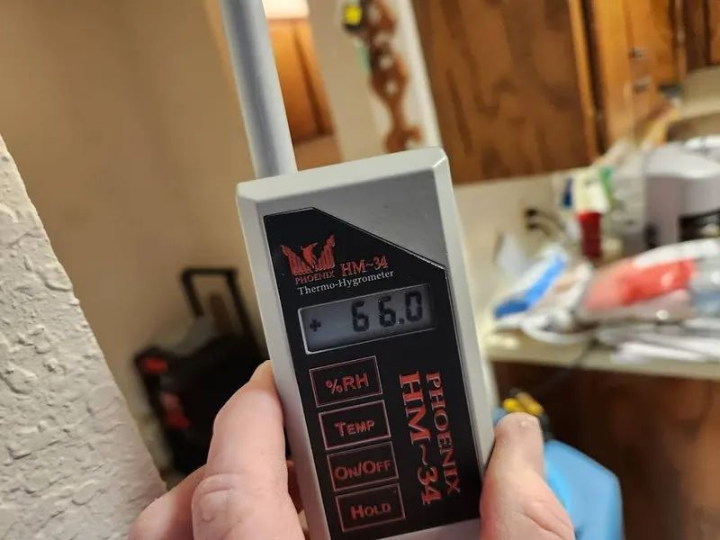 Hand holding a hygrometer reading 6.0 in a cluttered indoor room