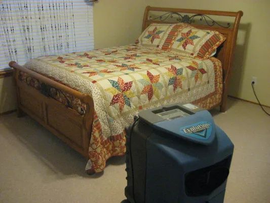 Bedroom with a wooden bed, patchwork quilt, and a blue floor fan in the foreground.