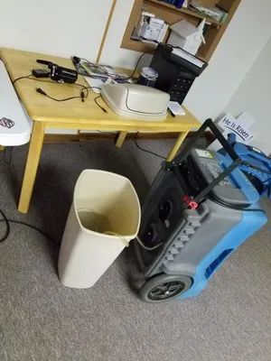 Office corner with a wooden desk, scattered electronics, a trash bin, and a blue industrial vacuum cleaner.