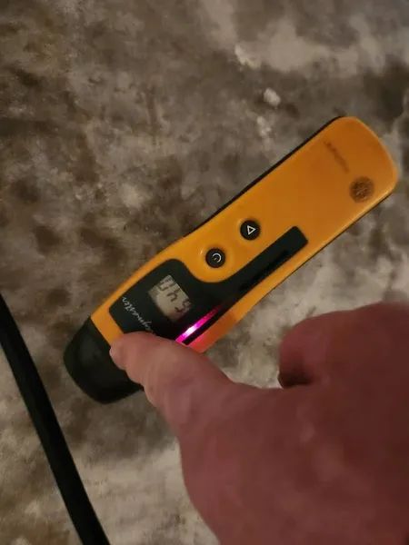 Hand holding a yellow handheld surface meter with black buttons and a small screen over a carpeted floor