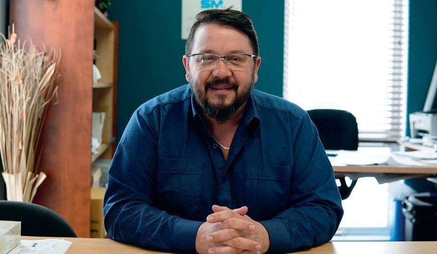 Un homme avec une barbe et des lunettes est assis à un bureau, les mains jointes.