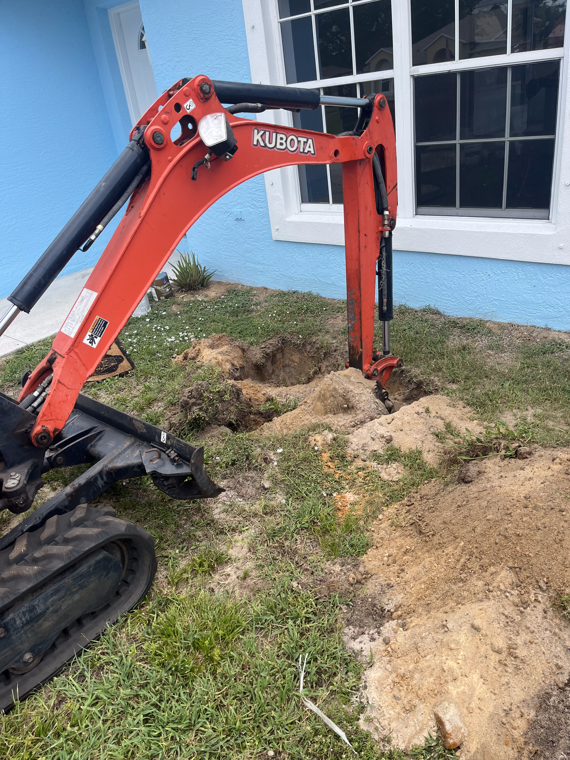 A small excavator is digging a hole in the grass in front of a house.