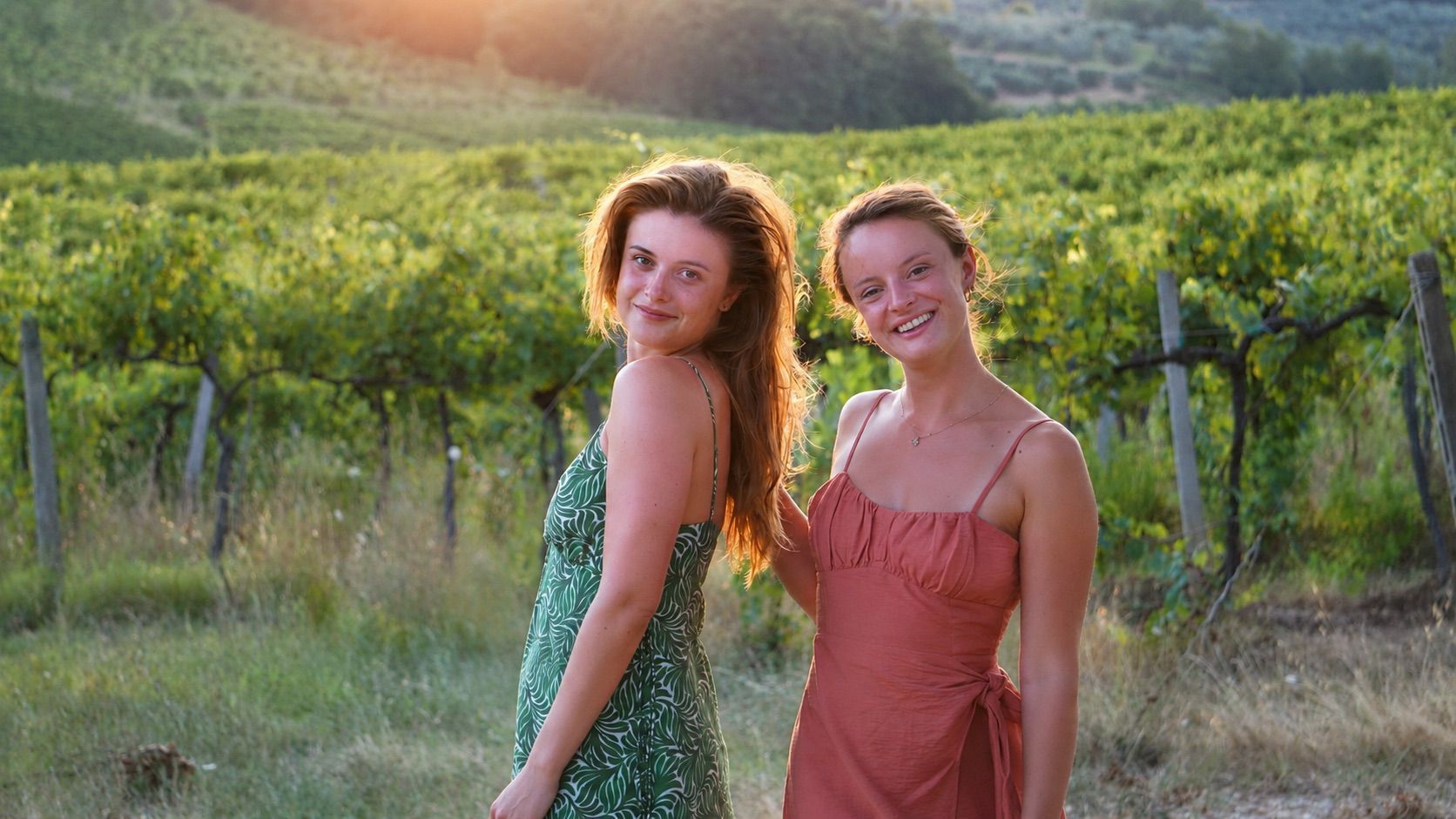 Twee vrouwen poseren in een zonovergoten wijngaard, met heuvels op de achtergrond.