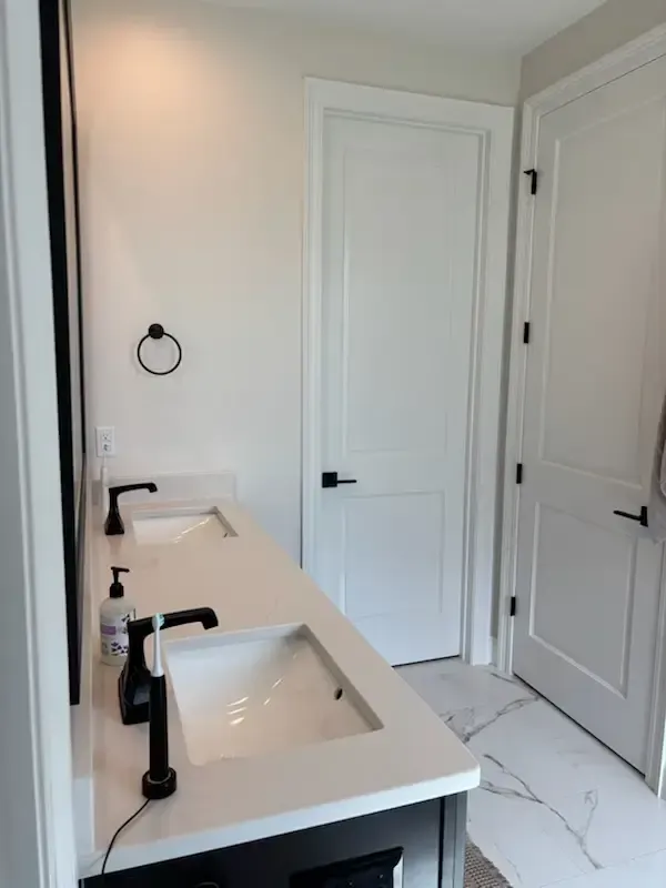 Modern bathroom with double sinks, black fixtures, white walls, and marble-look tile floor