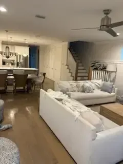 Open-plan living room with white sofas, wood floors, staircase, dining table, and kitchen in the background
