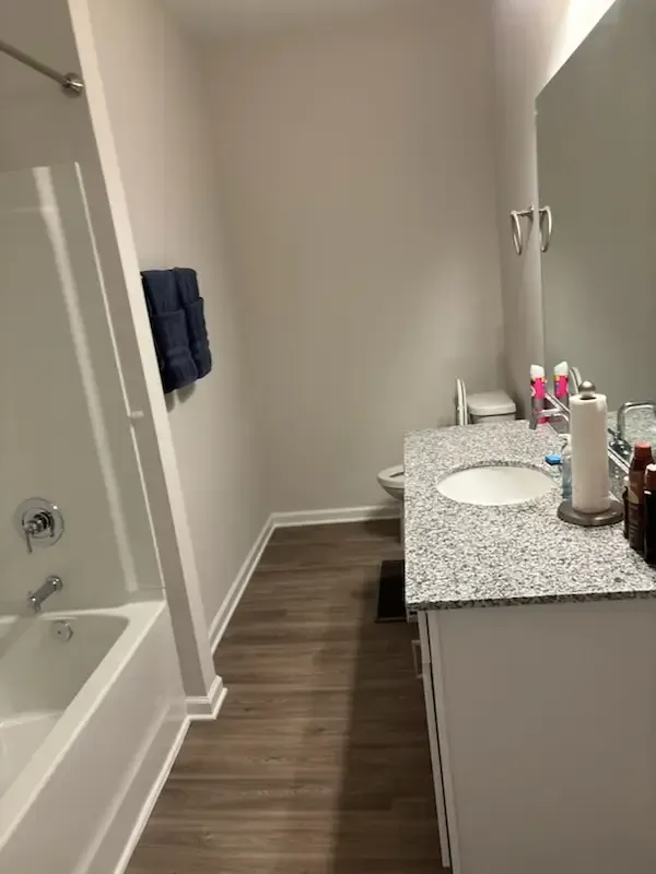 Small bathroom with bathtub, granite vanity, wood-look floor, and dark towel on wall.