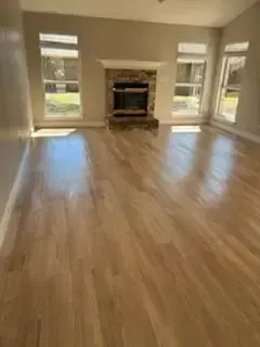 Empty living room with hardwood floors, large windows, and a fireplace.