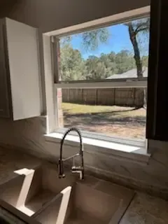 Kitchen sink under a large window overlooking a backyard with a fence and trees.