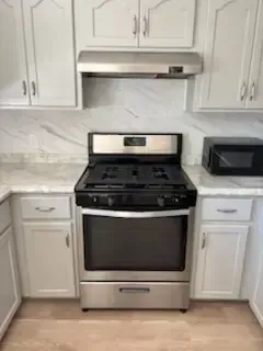 White kitchen with stainless steel gas stove, hood, and microwave between white cabinets and marble backsplash.