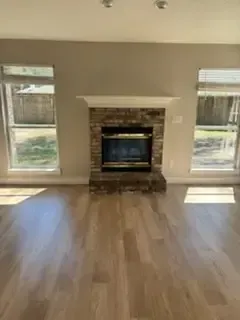 Empty living room with hardwood floors and a brick fireplace centered between two windows.