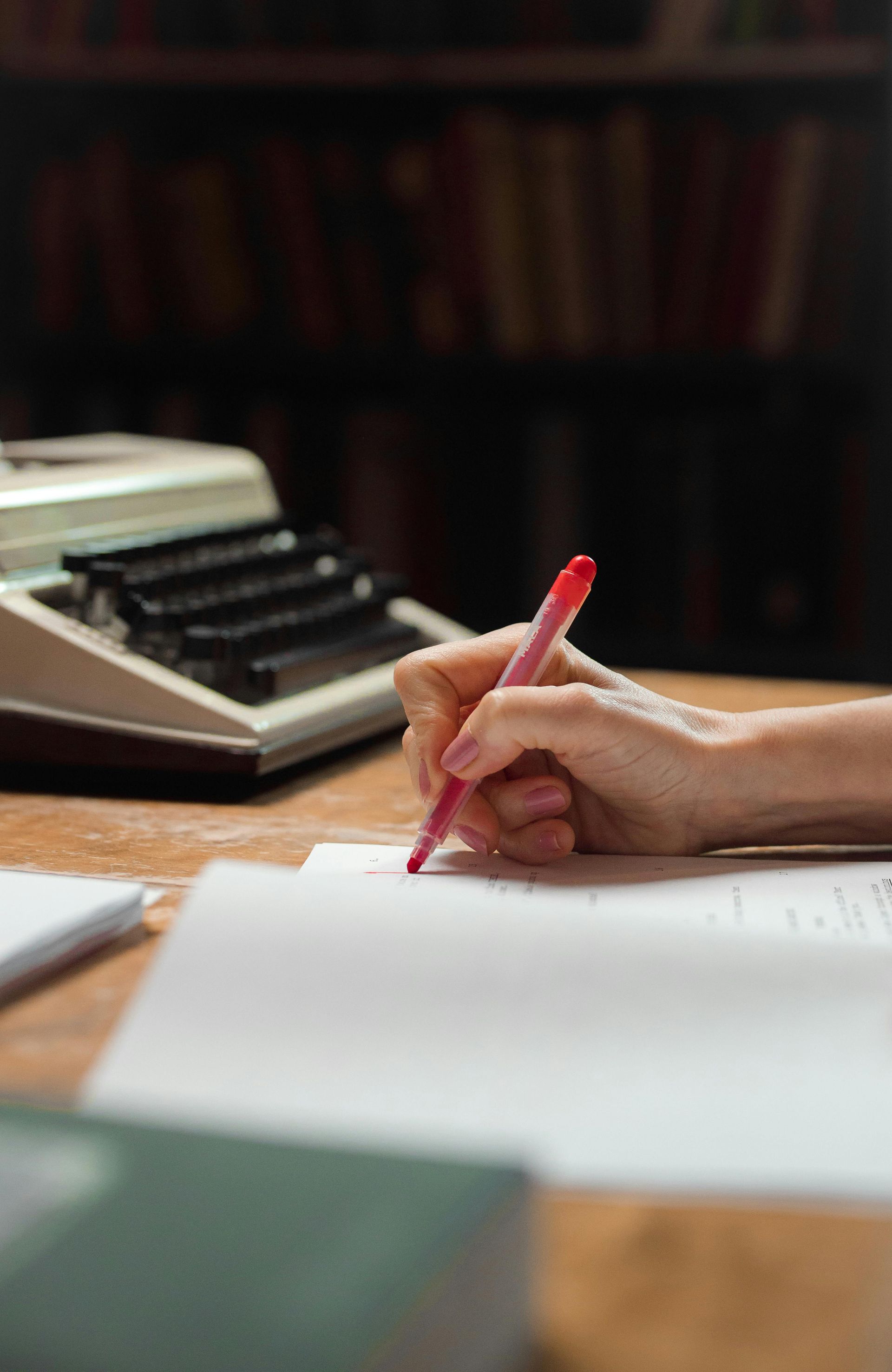 Person's hand writing with a red pen on paper at a desk, with a typewriter and bookshelf in the background.