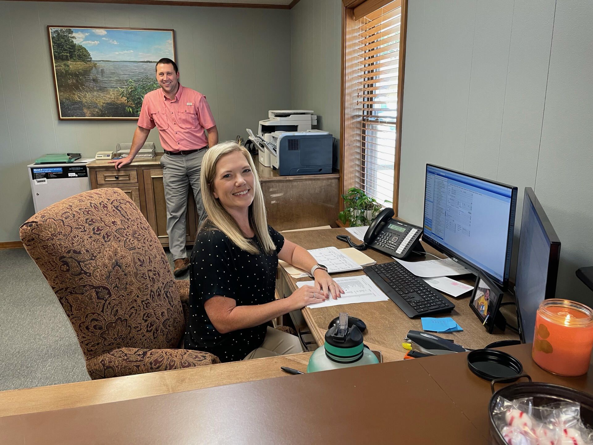 A woman is sitting at a desk in an office with a man standing behind her.