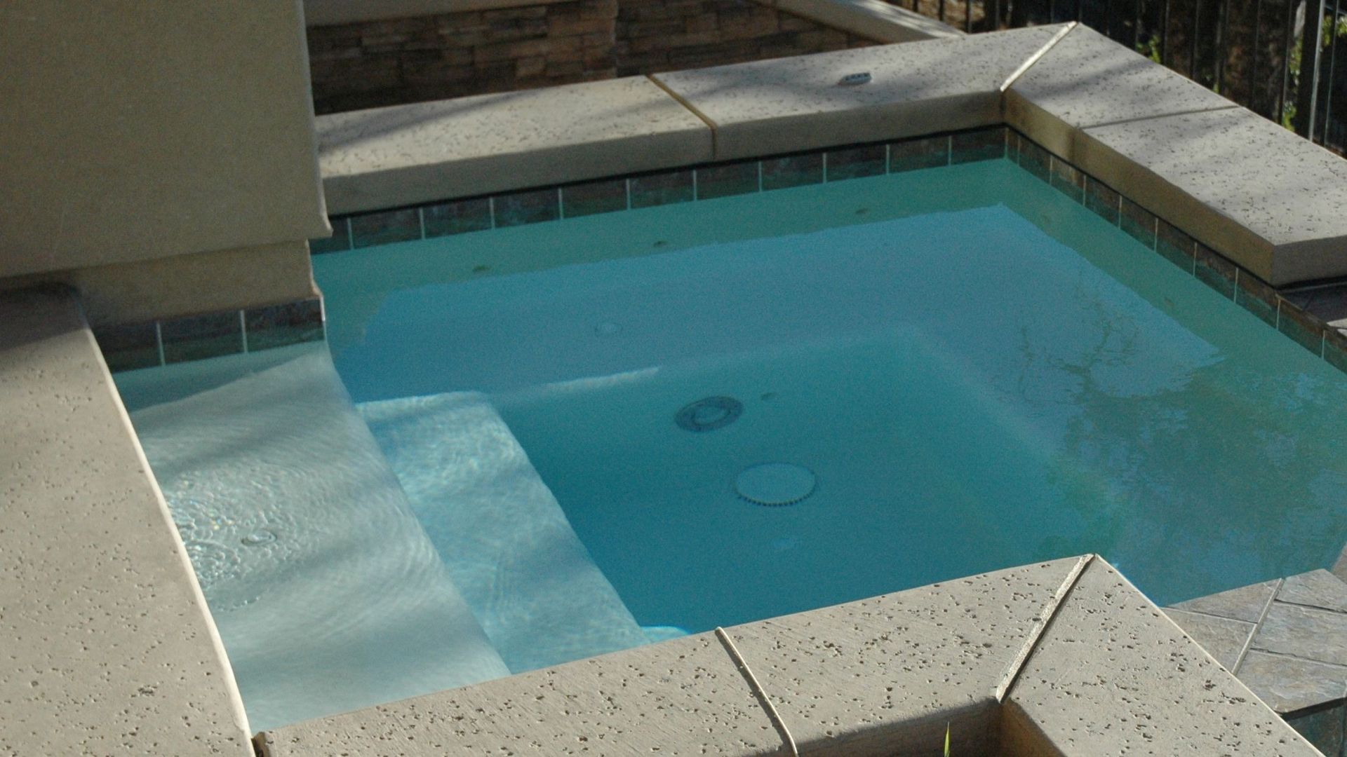 Small blue-tiled pool with a shallow ledge and steps beside a stone patio.