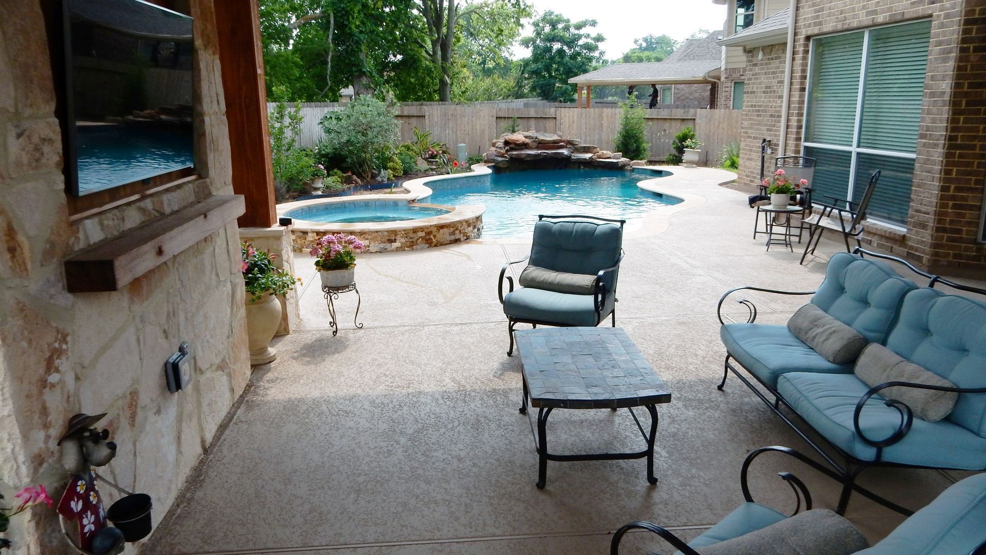 Backyard patio with blue pool, wicker chairs, and lounge seating beside a stone house