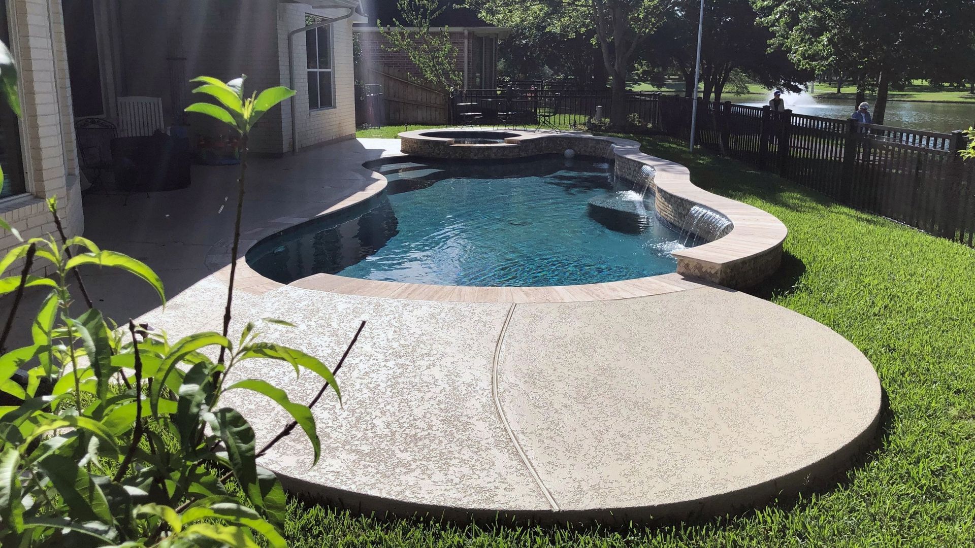 Backyard pool with a curved stone edge, concrete patio, and green lawn in bright sunlight