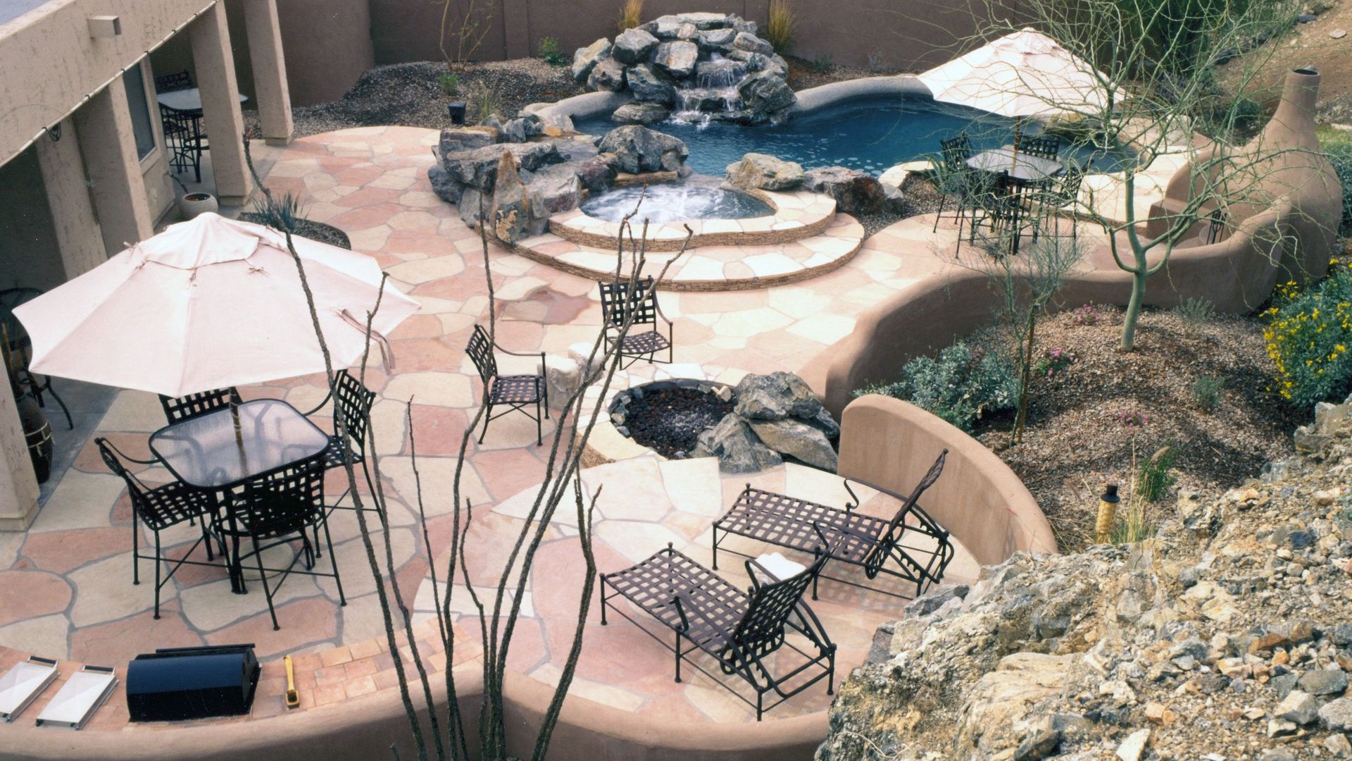 Backyard patio with round tables, white umbrellas, metal chairs, and a stone fire pit near a small pool